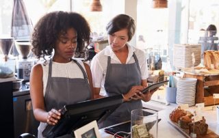 Ice cream shop new employee training shows two women in a food shop environment. One woman is showing the other how to operate the point of sale terminal. There's a tip jar on the counter.